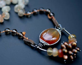 May include: A long necklace featuring a large carnelian gemstone pendant, surrounded by smaller beads in shades of brown, tan, and clear. The necklace is crafted with wire and leather accents.