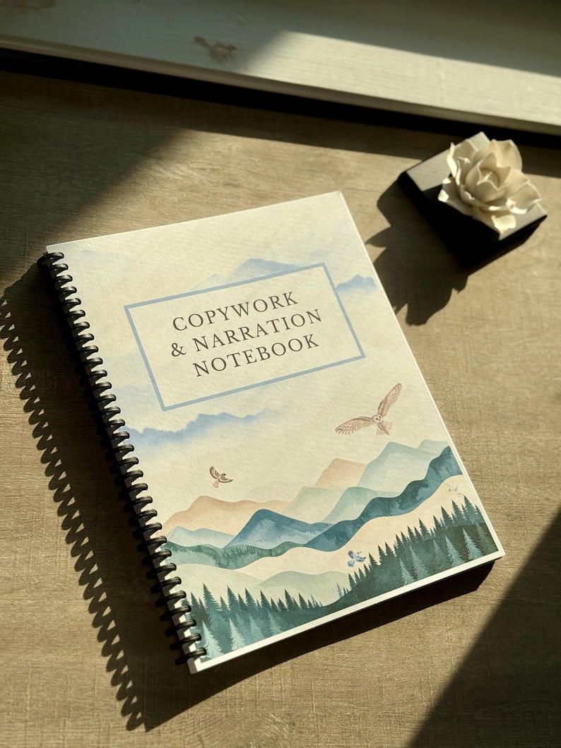 May include: Spiral-bound notebook with the text "COPYWORK & NARRATION NOTEBOOK" in a blue frame. The cover features a watercolor landscape of mountains, trees, and flying birds. The notebook is on a wooden surface.