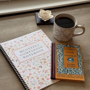 Puede incluir: Un cuaderno de espiral y un libro antiguo están junto a una taza de café. El cuaderno tiene un diseño floral y el texto "MCGUFFEY COMPANION NOTEBOOK". El libro antiguo se titula "MCGUFFEY'S ECLECTIC PRIMER". Una flor blanca está sobre un cuadrado negro.
