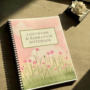 May include: A spiral-bound notebook with a cover featuring the text "COPYWORK & NARRATION NOTEBOOK" in a green-bordered box. The cover has a watercolor design of pink flowers and green grass against a pink sky. A white flower sits on a black square.