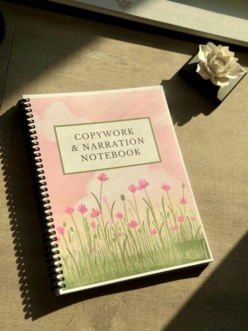 May include: A spiral-bound notebook with a cover featuring the text "COPYWORK & NARRATION NOTEBOOK" in a green-bordered box. The cover has a watercolor design of pink flowers and green grass against a pink sky. A white flower sits on a black square.