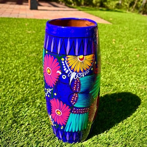 May include: A hand-painted, cylindrical ceramic vase with a vibrant blue background. The vase is adorned with colorful floral and bird designs in pink, yellow, green, and teal. The vase is standing on a green lawn.