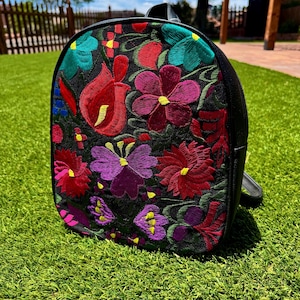 May include: A black backpack featuring a vibrant, multi-colored floral embroidery pattern on the front panel. The design includes various flower shapes in shades of red, purple, teal, and yellow, stitched directly onto the black fabric surface.