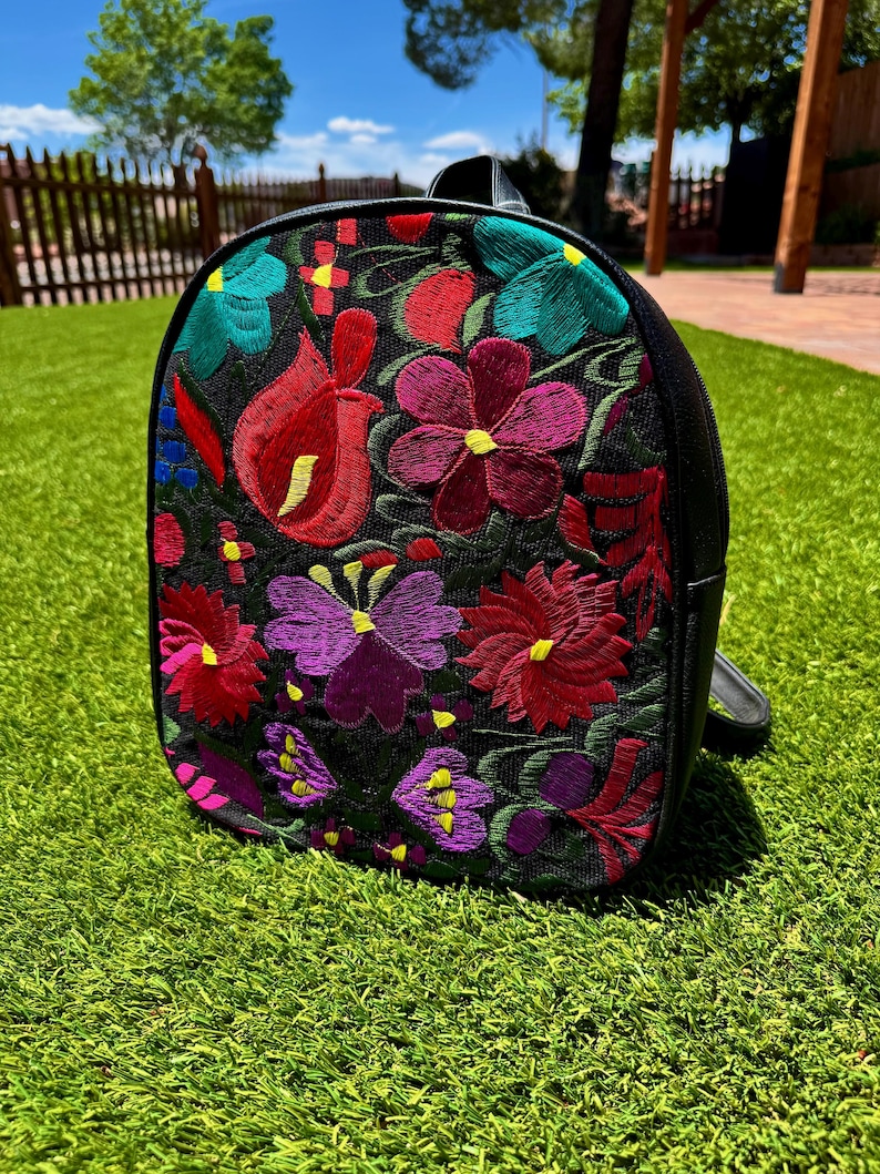 May include: A black backpack featuring a vibrant, multi-colored floral embroidery pattern on the front panel. The design includes various flower shapes in shades of red, purple, teal, and yellow, stitched directly onto the black fabric surface.