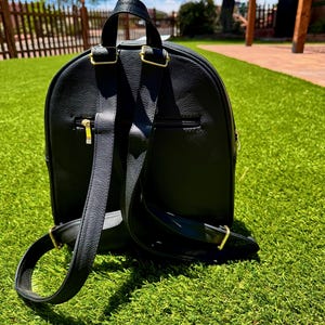 May include: A black backpack with a textured surface, featuring adjustable shoulder straps with gold-toned metal hardware, a top handle, and a horizontal zippered pocket on the back panel. The design includes a rounded top and a structured base.