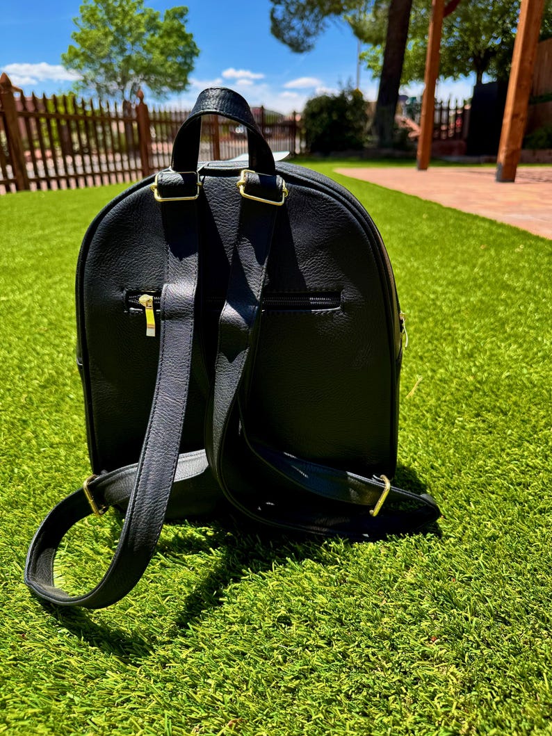 May include: A black backpack with a textured surface, featuring adjustable shoulder straps with gold-toned metal hardware, a top handle, and a horizontal zippered pocket on the back panel. The design includes a rounded top and a structured base.