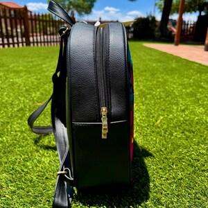 May include: A black backpack with a textured faux leather finish, featuring a side profile view. The bag includes a gold-toned metal zipper pull, adjustable shoulder straps, and a glimpse of a colorful, multi-patterned fabric panel on the front.