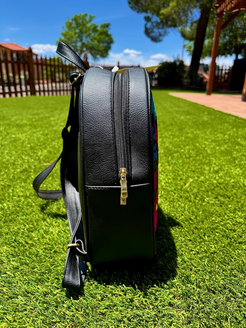 May include: A black backpack with a textured faux leather finish, featuring a side profile view. The bag includes a gold-toned metal zipper pull, adjustable shoulder straps, and a glimpse of a colorful, multi-patterned fabric panel on the front.