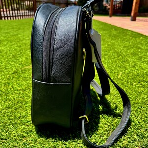 May include: A black backpack with a textured surface, featuring a side zipper compartment, a top handle, and adjustable shoulder straps. The bag includes a white tag attached to the top handle and gold-toned metal hardware on the strap attachments.