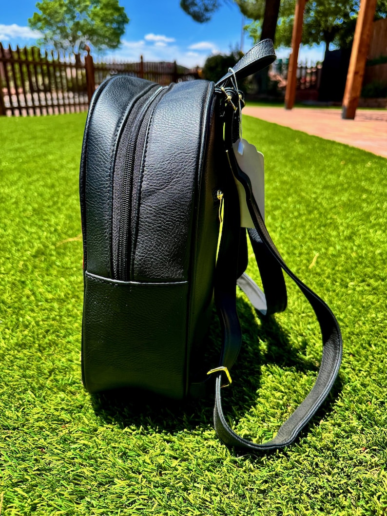 May include: A black backpack with a textured surface, featuring a side zipper compartment, a top handle, and adjustable shoulder straps. The bag includes a white tag attached to the top handle and gold-toned metal hardware on the strap attachments.