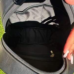 May include: A black backpack with a textured exterior and a smooth black fabric interior lining. The main compartment features a zippered pocket with a gold-toned metal pull and an open slip pocket along the back wall for organized storage.