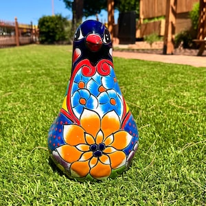 以下が含まれることがあります： 鮮やかにペイントされたセラミック製の鳥の置物。鳥は、青い頭、赤い嘴、オレンジと青の花柄で飾られた体を持つ光沢のある仕上がりです。装飾品は緑の草の上に置かれています。