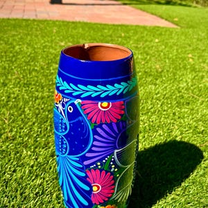 May include: A hand-painted, cylindrical ceramic vase with a vibrant blue base. The vase features a detailed design of a bird and flowers in shades of pink, purple, and turquoise. The interior is a warm brown color.