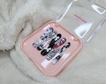 May include: A collection of press-on nails in a pink case. The nails display diverse black and white designs, including swirls, floral patterns, and abstract art. Some nails have red accents. The case has the text "Homemade Nails".