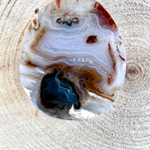 May include: A polished, oval-shaped pendant with a silver bail. The stone features a natural agate pattern with layers of white, brown, and red hues. A dark blue and black area is visible near the bottom of the pendant.