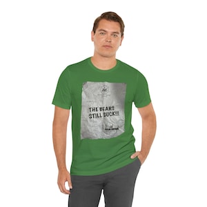 May include: Green t-shirt with a graphic on the front that reads "THE BEARS STILL SUCK!!!" in bold black letters. The graphic is designed to look like a crumpled piece of paper with a seal and signature.
