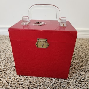 Könnte beinhalten: Eine rote Vintage-Box mit einem durchsichtigen Kunststoffgriff und einem messingfarbenen Verschluss. Die Box ist quadratisch und scheint aus einem strukturierten Material zu bestehen. Der Griff ist gewölbt und an der Oberseite der Box befestigt.