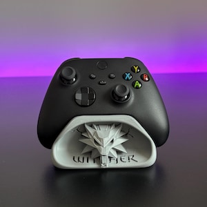 May include: A black wireless game controller resting on a light grey stand. The stand features a sculpted wolf head emblem and the text WITCHER embossed on the front. The controller has standard black joysticks, a directional pad, and coloured action buttons.