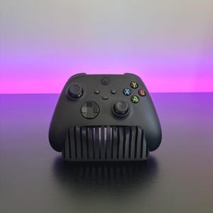 Può includere: Un controller per videogiochi wireless nero opaco appoggiato su un supporto a lamelle nero. Il controller presenta due levette analogiche, un pad direzionale e quattro pulsanti azione contrassegnati dalle lettere A, B, X e Y in verde, rosso, blu e giallo.