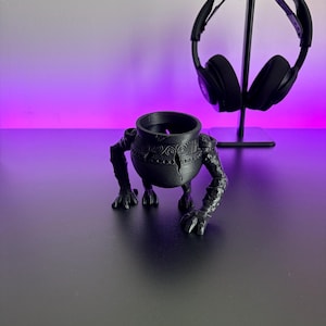 May include: A matte black 3D printed planter shaped like a pot with three textured, clawed limbs extending outwards. The central pot features a circular opening at the top and intricate, carved spiral patterns around the exterior surface.
