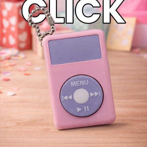 May include: A pink MP3 player keychain with a purple screen and control buttons. The text "GIFT A CLICK" is above the player. The background includes gift boxes and confetti on a wooden surface.