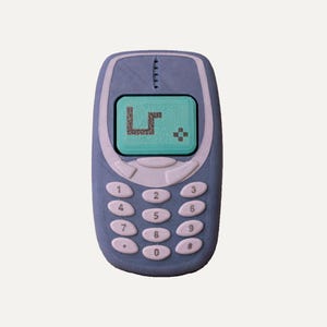 Può includere: Una replica di telefono cellulare blu con uno schermo verde chiaro che mostra un gioco di serpente pixelato scuro. Il dispositivo include un tastierino numerico standard con pulsanti ovali bianchi e una griglia dell'altoparlante.