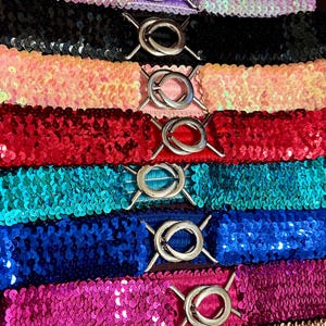 Könnte beinhalten: Eine Sammlung von Pailletten-Gürteln in verschiedenen Farben, darunter Rosa, Schwarz, Rot und Blau. Jeder Gürtel hat einen silberfarbenen Verschluss. Die Gürtel sind horizontal angeordnet und zeigen ihre schimmernde Textur und ihr Design.