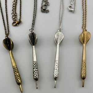Può includere: Una collezione di collane con ciondoli a forma di freccette in oro, argento e bronzo. Ogni collana presenta un ciondolo a freccetta dettagliato sospeso a una catena abbinata. Le freccette hanno manici strutturati e alette a forma di piuma.