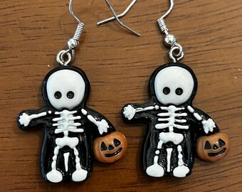 Puede incluir: Un par de pendientes con temática de Halloween. Cada pendiente presenta una figura de esqueleto con túnica negra con calavera y caja torácica blancas, sosteniendo una calabaza naranja. Los pendientes tienen cierres de gancho plateados.