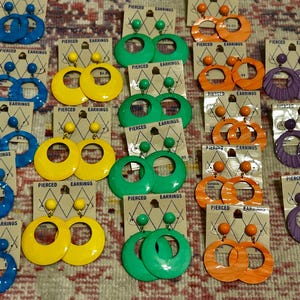 May include: A collection of vintage pierced earrings in various colors, including blue, yellow, green, orange, and purple. Each pair is displayed on a card with the words "PIERCED EARRINGS". The earrings have a large hoop design.