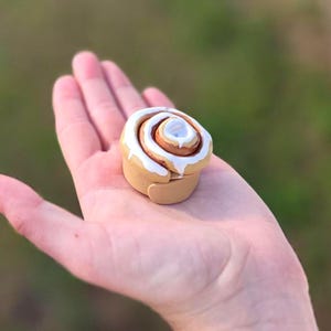 May include: A miniature cinnamon roll figurine crafted from tan, brown, and white material. The spiral-shaped pastry features a smooth, light tan base with a coiled brown center and a thick, glossy white icing drizzle dripping down the sides.