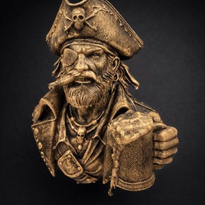 May include: A bronze-colored pirate bust with a detailed design. The pirate wears a tricorn hat with a skull and crossbones, an eye patch, and a jacket. He holds a mug of beer and a cigar. The bust is set against a black background.