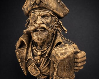 May include: A bronze-colored pirate bust with a detailed design. The pirate wears a tricorn hat with a skull and crossbones, an eye patch, and a jacket. He holds a mug of beer and a cigar. The bust is set against a black background.