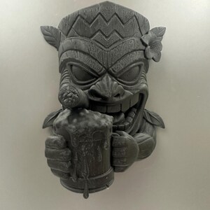 May include: A dark gray tiki mask sculpture with a smiling face, holding a mug of beer and a cigar. The mask has detailed carvings, including a floral accent and leaf details. The beer mug has dripping foam.