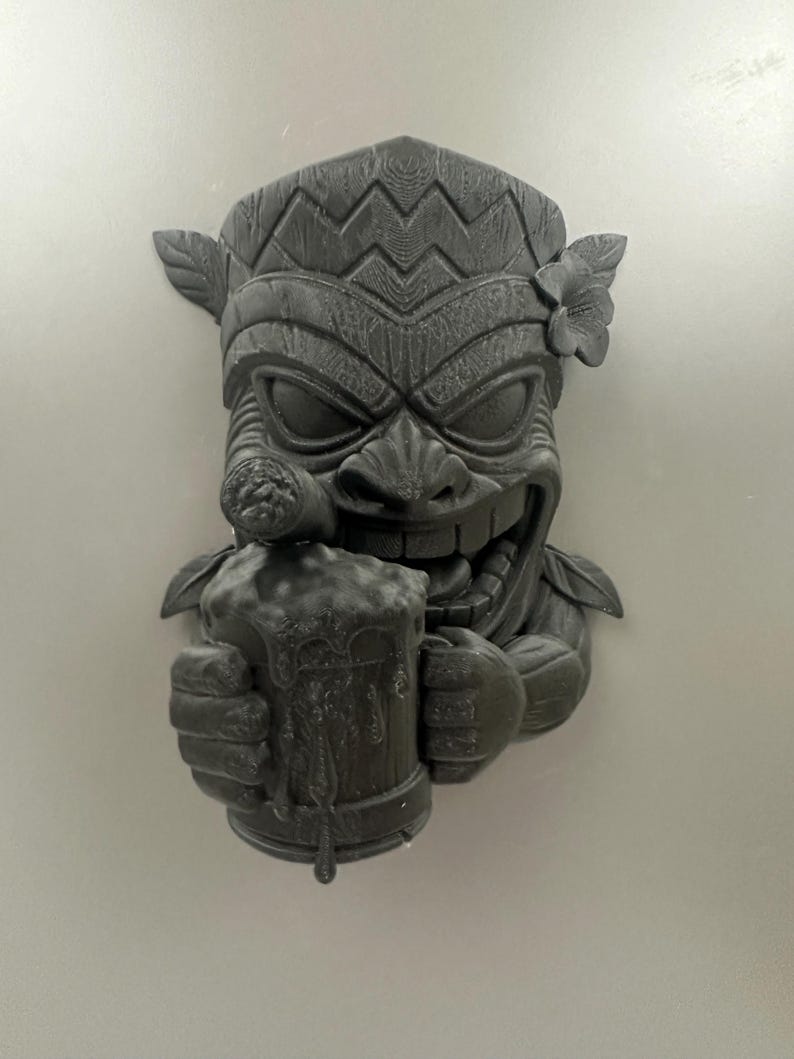 May include: A dark gray tiki mask sculpture with a smiling face, holding a mug of beer and a cigar. The mask has detailed carvings, including a floral accent and leaf details. The beer mug has dripping foam.