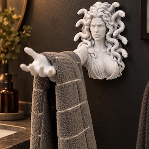 May include: A wall-mounted towel holder sculpted in the likeness of Medusa with snakes for hair. The figure is light gray with a textured stone-like finish, featuring an outstretched arm that functions as a rack for a gray textured towel.