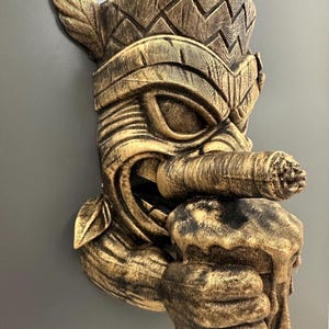 May include: A detailed, golden-brown tiki mask with a cigar in its mouth and a mug of beer in its hand. The mask has intricate carvings and a weathered appearance, suggesting a vintage or tropical theme. The mask is mounted on a gray surface.