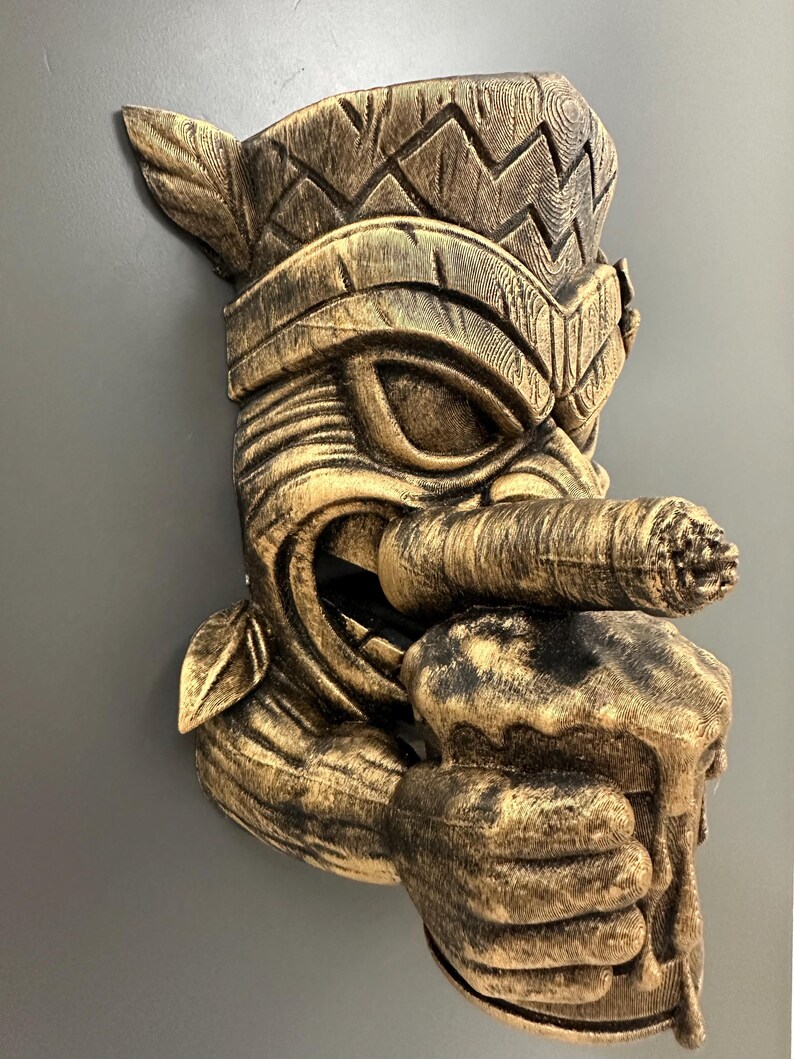 May include: A detailed, golden-brown tiki mask with a cigar in its mouth and a mug of beer in its hand. The mask has intricate carvings and a weathered appearance, suggesting a vintage or tropical theme. The mask is mounted on a gray surface.