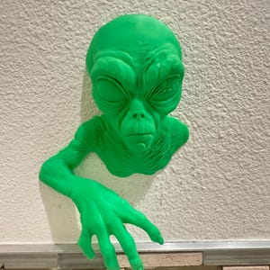 Puede incluir: Escultura de una cabeza y un brazo de alienígena verde montada en una pared blanca texturizada. El alienígena tiene ojos grandes, una nariz pequeña y una mano extendida. La escultura es de color verde vibrante.