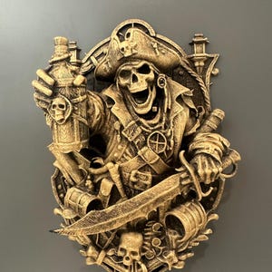 Puede incluir: Decoración de pared de calavera pirata dorada. La calavera lleva un sombrero y una chaqueta, sosteniendo una botella y una espada. El diseño incluye un escudo, un barril y otros elementos temáticos piratas. La decoración de pared está montada sobre una superficie gris.