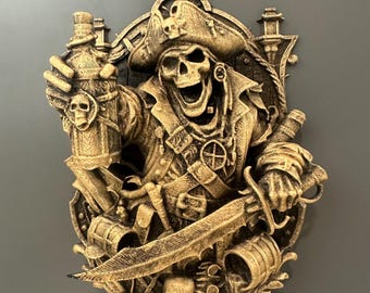 May include: A gold-colored pirate skull wall decor. The skull wears a hat and jacket, holding a bottle and sword. The design includes a shield, barrel, and other pirate-themed elements. The wall decor is mounted on a gray surface.