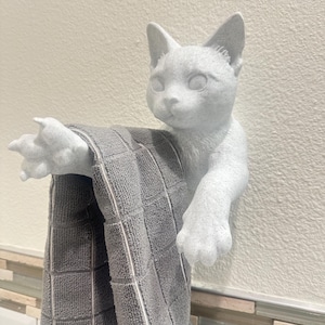 Può includere: Un gancio da parete a forma di gatto bianco che tiene un asciugamano grigio. Il gatto ha un viso e delle zampe dettagliate, che sembrano allungarsi. L'asciugamano è grigio con un motivo a quadri e bordi bianchi.
