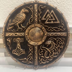 May include: A round, black and gold shield featuring Norse symbols. The shield has a central boss and is divided into quadrants with a raven, a symbol of Thor's hammer, a serpent, and the Valknut symbol. The shield is decorated with intricate knotwork.