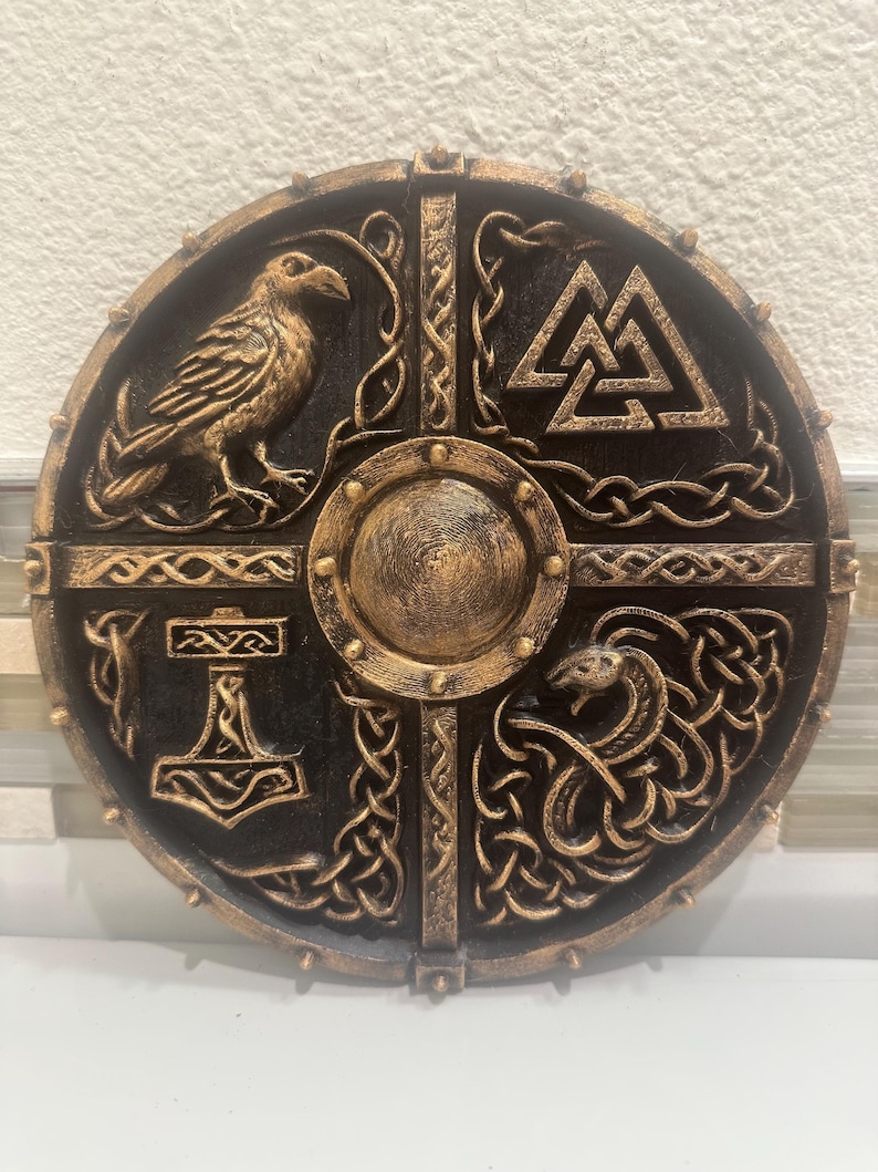 May include: A round, black and gold shield featuring Norse symbols. The shield has a central boss and is divided into quadrants with a raven, a symbol of Thor's hammer, a serpent, and the Valknut symbol. The shield is decorated with intricate knotwork.