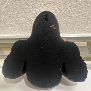 May include: A black wall hanging with a textured surface. The wall decor has a rounded top and two side extensions, resembling a stylized figure. A small hole is visible at the top for hanging.