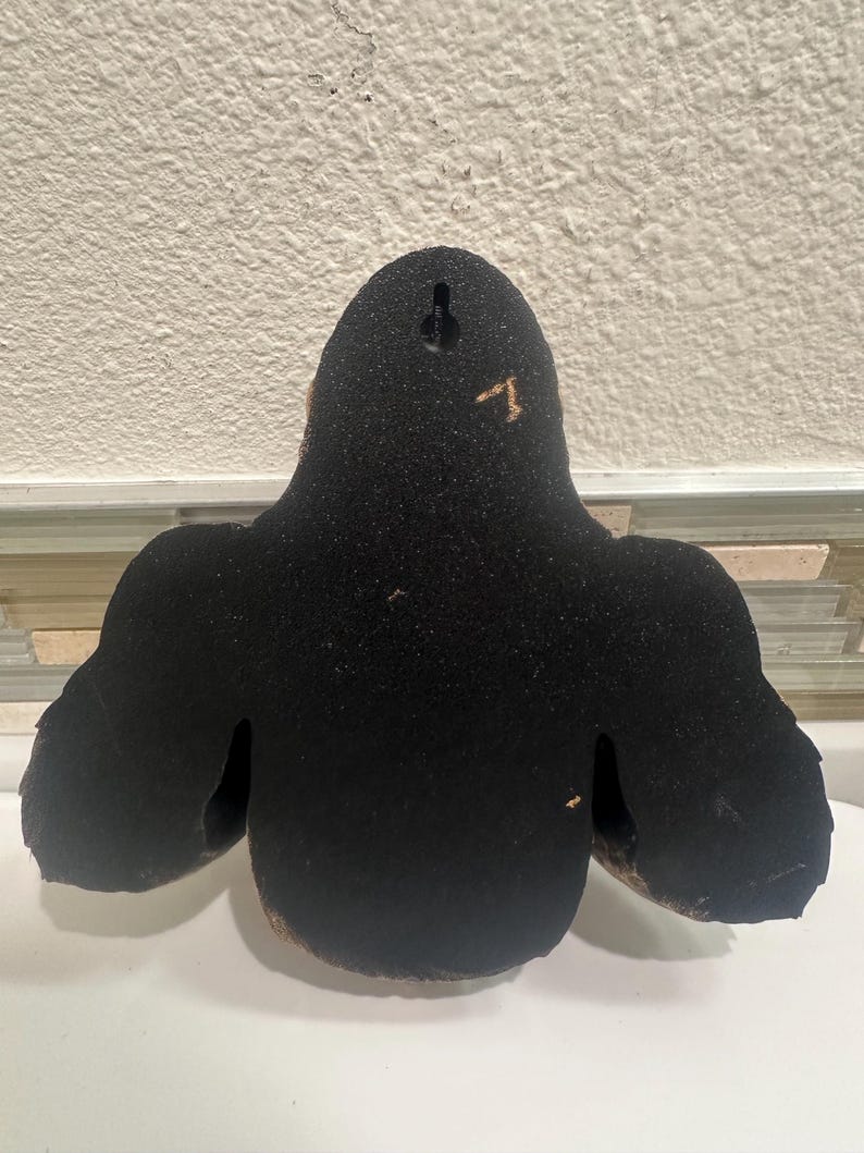 May include: A black wall hanging with a textured surface. The wall decor has a rounded top and two side extensions, resembling a stylized figure. A small hole is visible at the top for hanging.