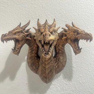 May include: A detailed, three-headed dragon sculpture in shades of brown, mounted on a white wall. Each dragon head has open jaws, sharp teeth, and textured scales. The sculpture is a decorative piece, likely for home decor.