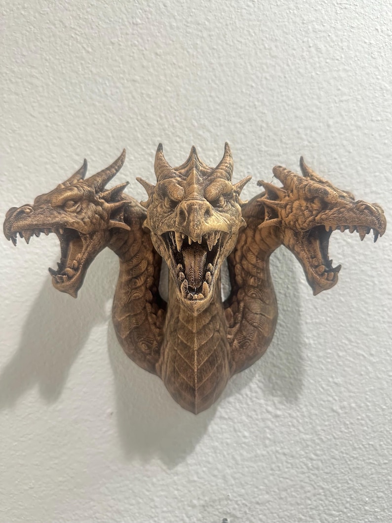 May include: A detailed, three-headed dragon sculpture in shades of brown, mounted on a white wall. Each dragon head has open jaws, sharp teeth, and textured scales. The sculpture is a decorative piece, likely for home decor.