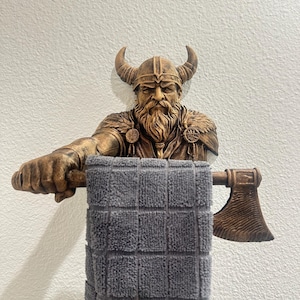Puede incluir: Un toallero con forma de figura vikinga de color bronce, con un casco con cuernos y un hacha. El toallero sostiene una toalla gris a cuadros. El vikingo tiene una larga barba y está montado en una pared blanca.