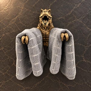 May include: A gold-colored T-Rex dinosaur wall-mounted towel holder featuring a detailed sculpted head with an open mouth and two clawed hands extending forward to grip and hold a folded gray textured towel in place on the wall.